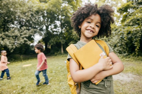 young student holding notebook and smiling as other kids play in the background