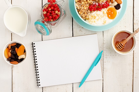 blank notebook with pen laying on a table with fruit, milk, honey and a yogurt bowl