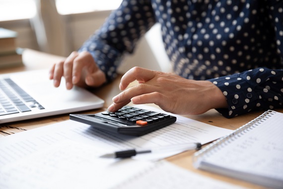 woman using a calculator and laptop with notebook and papers on a desk