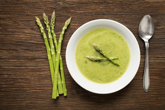 bowl of asparagus soup on a table with fresh asparagus and a spoon